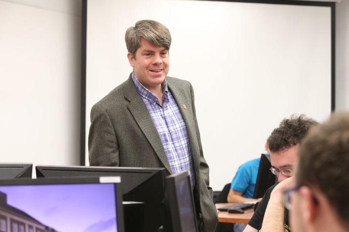 A man wearing a purple plaid shirt and a grey suit jacket stands at the front of a computer lab/classroom. Seats in the room are filled by students.