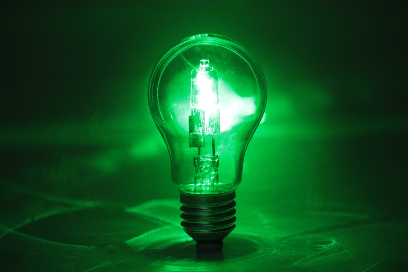 A transparent lightbulb is pictured on a green background. A laser pointed at the bulb from behind makes it appear to be lit. Reflections from the bulb's glass bounce around the background.